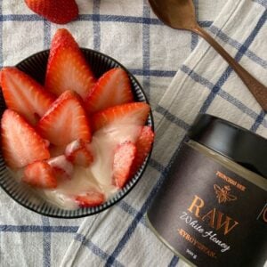 a jar of raw sainfoin white honey next to a bowl with a strawberry